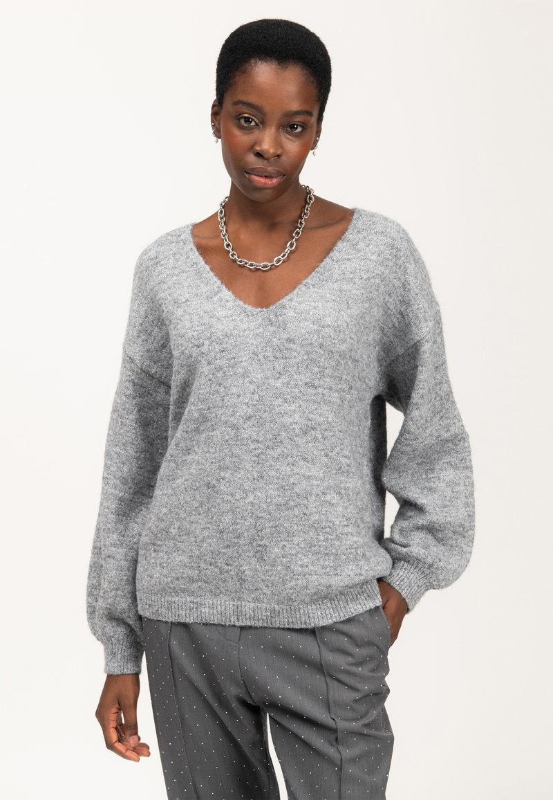 Donna che indossa un ampio maglione grigio a scollatura a V, una collana a catena e pantaloni grigi a righe sottili con pois bianchi, in posa con una mano in tasca.