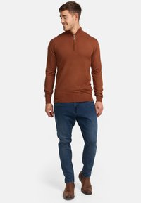 Brauner gestrickter Zip-Up-Pullover, taillierter Schnitt, lange Ärmel, gerippte Bündchen und Saum. Kombiniert mit blauen Jeans und braunen Ankle-Boots.