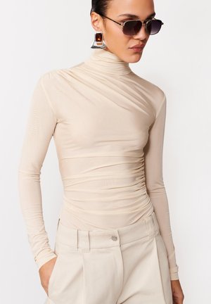 Femeie purtând un pulover bej cu guler înalt și pliuri, pantaloni cu talie înaltă, cercei triunghiulari din chihlimbar și argint, și ochelari de soare cu ramă cat-eye, pozând pe un fundal alb.