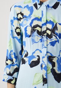Blomstrete skjorte med blå nyanser og lys blå bakgrunn, med krage og trekvartlengde ermer. Blomstene er i nyanser av blått, sort og grønt.