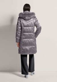 Vrouw in een lange, glanzende grijze pufferjas met een met bont afgewerkte capuchon, zwarte wijde broek en lichtgrijze hakken, staand met haar rug naar de kijker.