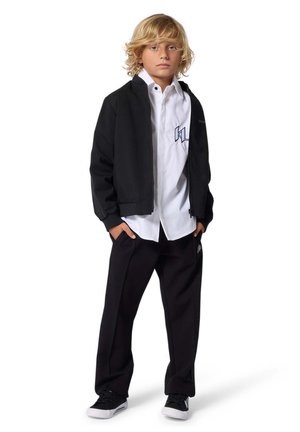 Schwarze Bomberjacke über einem weißen Hemd, kombiniert mit schwarzen Hosen und schwarzen Turnschuhen. Mit Logo auf Hemd und Hose, lässige Passform.