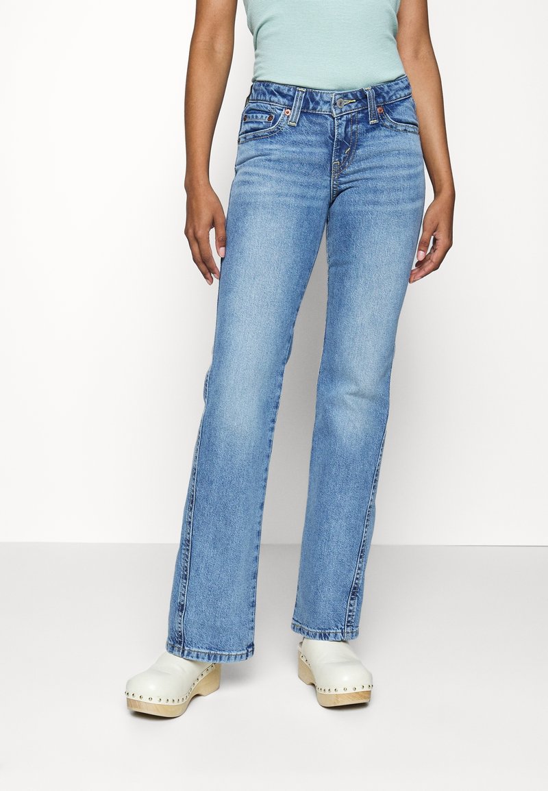 Personne portant un jean droit bleu clair, un haut vert menthe et des sabots blancs en bois à plateforme avec des clous métalliques.