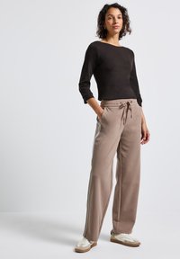 Mujer de pie con las manos en los bolsillos, vistiendo una camiseta de manga larga negra, pantalones anchos de tiro alto beiges con cordón y rayas laterales, y zapatillas blancas.