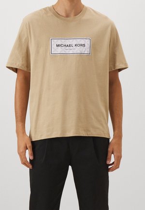 Beżowa bawełniana koszulka z krótkim rękawem o luźnym kroju, z prostokątną białą metką z napisem "MICHAEL KORS" oraz "NEW YORK CITY" w czarnym kolorze.
