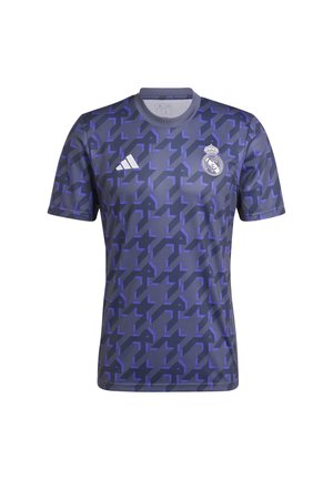 Mann trägt ein dunkelblaues und lila Real Madrid-Shirt mit geometrischem Muster und marineblaue Adidas-Hose und steht vor weißem Hintergrund.