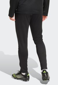 Sorte atletikbukser med slank pasform, glat tekstur og kontrasterende hvidt logo. Et par sorte spikessko med grønne accenter synlige ved anklen.