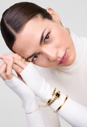 Les bijoux dorés comprennent une montre avec un cadran noir, deux bracelets avec des pierres, et un bracelet délicat en chaîne au poignet d'un mannequin.