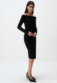 Abito nero scollato sulle spalle con maniche lunghe, design aderente, scollatura a coste e orlo fino al ginocchio. Indossato con tacchi a rocchetto neri.