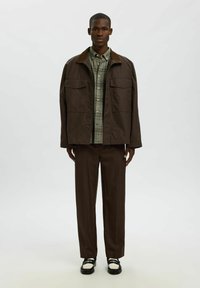 Rjav Oversized plašč z bež ovratnikom, nošen čez zeleno karirasto srajco, v kombinaciji z rjavimi hlačami in črno-belimi čevlji.