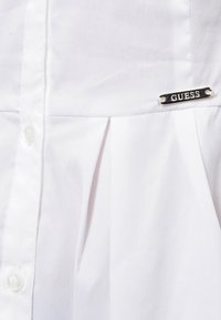 Chemise blanche boutonnée avec un design plissé, arborant un accent logo argenté "GUESS" sur la poitrine et une texture lisse semblable au coton.
