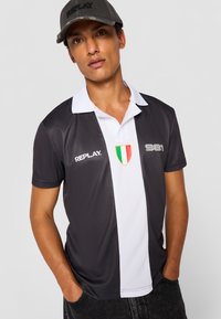Sort-hvid kortærmet polo med krave, der har et farverigt Italien badge og teksten "REPLAY." trykt i hvid. Tekstureret stof.
