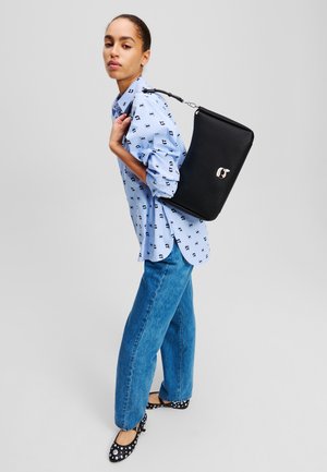 Sac à bandoulière en cuir noir avec une texture lisse, de forme rectangulaire et des accents métalliques. Porté sur le dos avec une chemise à rayures bleues et un jean.