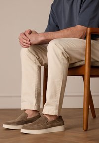 Suede slip-on shoes in brown with a smooth texture, featuring a white rubber sole. Worn with light beige pants, seated on a wooden chair.