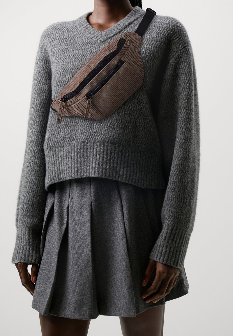 Personne portant un pull gris texturé et une jupe plissée grise avec un sac bandoulière en velours côtelé marron avec deux fermetures éclair noires.