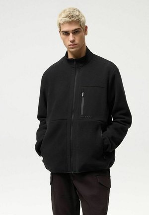 Młody mężczyzna o krótkich blond włosach, ubrany w czarną polarową kurtkę z zasuwaną kieszenią na piersi i rękami w bocznych kieszeniach, stojący na jednolitym tle.