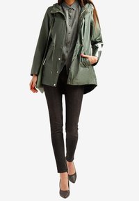 Wodoszczelna zielona kurtka z kapturem, zapięcie na guziki i zamek błyskawiczny, kieszenie boczne; w zestawie z ciemnymi dżinsami i szarymi butami na wysokim obcasie.
