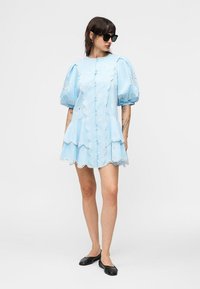 Γυναίκα που φοράει μίνι φόρεμα με φουσκωτά μανίκια σε ανοιχτό μπλε χρώμα με κυματιστές άκρες, κουμπιά μπροστά, μαύρα χαμηλά παπούτσια και μαύρα γυαλιά ηλίου, στέκεται μπροστά σε λευκό φόντο.