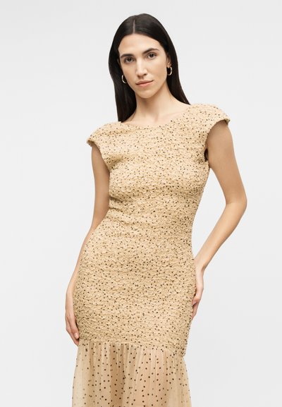 Γυναίκα με μακριά σκούρα μαλλιά, ντυμένη με μια μπεζ υφασμάτινη φόρεμα με τούλινη φούστα που έχει πουά σχεδιασμό, ποζάρει μπροστά σε ένα απλό φωτεινό φόντο.