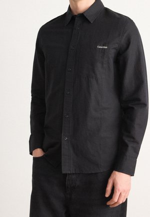 Mann trägt ein schwarzes, langärmliges Hemd mit Brusttasche und Calvin Klein Logo-Patch auf der linken Seite.