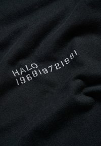Schwarzer Baumwollstoff mit dem Text "HALO" und den Zahlen "196819721981" in Weiß. Glatte Textur mit subtilen Falten.