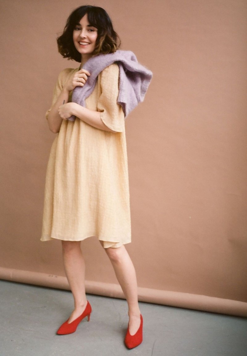 Keltainen, teksturoitu mekko, jossa on löysät hihat, kantaa vaalean lilaa neuletta olalla. Punaiset kärkitossut täydentävät asukokonaisuuden.