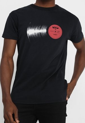 Homme portant un T-shirt noir avec un graphisme d'un disque vinyle partiellement visible et une étiquette rouge sur la poitrine.