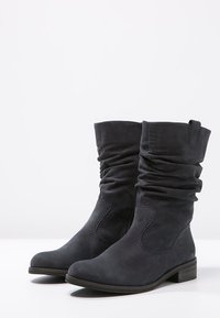 Botas de ante negras hasta la mitad de la pantorrilla con cañas fruncidas, tacones bajos apilados, punteras redondeadas y cierres de cremallera lateral sobre un fondo blanco.