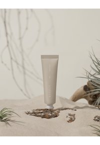 Beige presskammare med vit text, med en texturerad bas, placerad på sand med sparsamma gröna växter och drivved i bakgrunden.