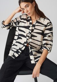 Camicia button-up beige con stampa astratta nera, caratterizzata da una vestibilità rilassata e maniche arrotolate. Indossata con pantaloni neri, in posa seduta.