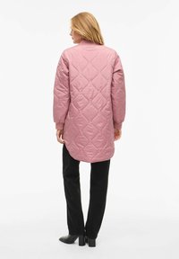 Prešita plašč z dolgimi rokavi v mehki roza barvi, z vzorcem diamantov, zaobljenim ovratnikom in daljšim zadnjim delom, kombiniran s črnimi hlačami in škornji z visokimi petami.