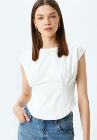 Witte, aangesloten blouse met kapmouwen, plooien aan de voorkant en een gladde textuur, gecombineerd met hoogsluitende blauwe jeans.