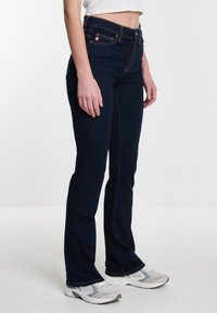 Jeans de mezclilla azul oscuro de corte ajustado, con costuras amarillas y bolsillos estándar. Combinados con zapatillas deportivas blancas y grises.