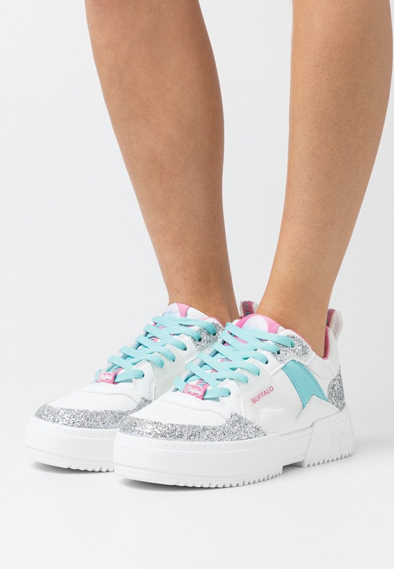 Baskets blanches avec accents en paillettes argentées, lacets bleus et détails roses. Dotées d'une semelle épaisse et du logo de la marque sur le côté.