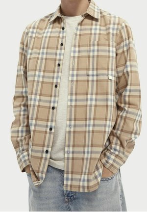 Mand iført beige og blå ternet skjorte med knapper over hvid t-shirt med hænderne i lommerne på lyseblå jeans.
