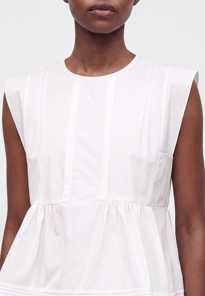 Femme portant un chemisier blanc sans manches avec des plis verticaux et un détail de taille ample et froncé sur un fond clair.