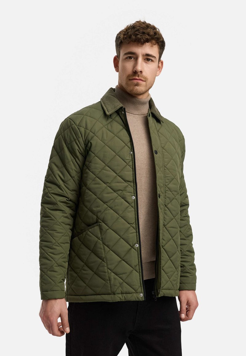 Quiltet grønn jakke med cordfløyelskrage, har frontglidelås, trykknapper og to sidelommer. Båret over en beige genser.