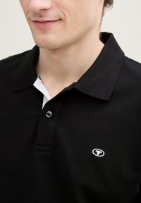 Svart polo skjorta med vit krage, som har en enstaka knapp och en liten broderad logotyp på vänster bröst. Tillverkad av bomull, med texturerad yta.
