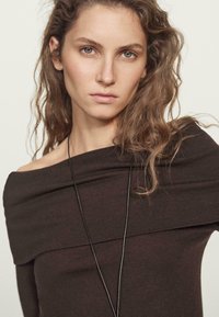 Brauner schulterfreier Oberteil aus weichem Stoff, mit weitem Ausschnitt und subtilen Raffung. Eine dünne schwarze Halskette sorgt für einen Kontrast.