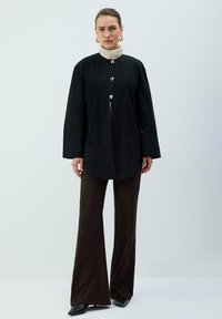 Top nero a maniche lunghe con bottoni dorati e un colletto alto bianco, abbinato a pantaloni marroni a gamba larga. Tessuto liscio e design minimale.