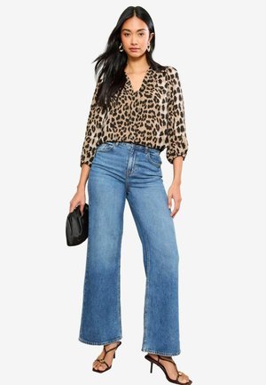 Blouse imprimée léopard avec un décolleté en V, associée à un jean large bleu clair. Le mannequin tient une pochette noire, portant des talons avec des brides.