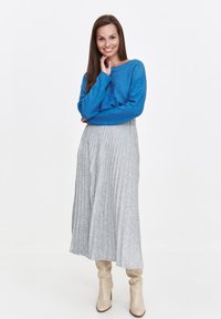Niebieski sweter z dzianiny z długimi rękawami, zestawiony z szarym plisowanym spódnicą. Nosi beżowe, luźne botki, stojąc na białym tle.