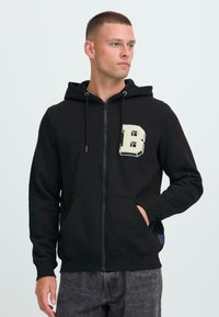 Uomo che indossa una felpa nera con zip, con una grande "B" color crema sul petto e le mani nella tasca frontale, che guarda verso destra.
