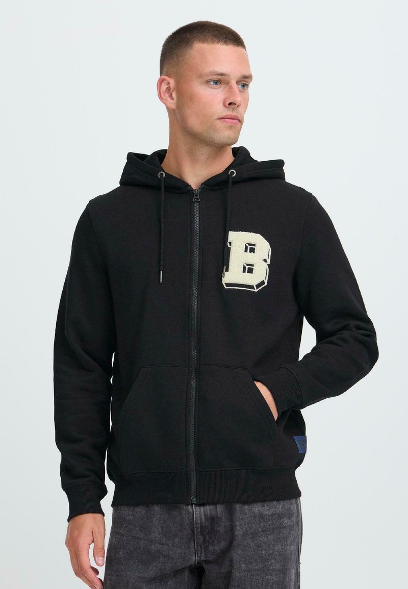 Uomo che indossa una felpa nera con zip, con una grande "B" color crema sul petto e le mani nella tasca frontale, che guarda verso destra.