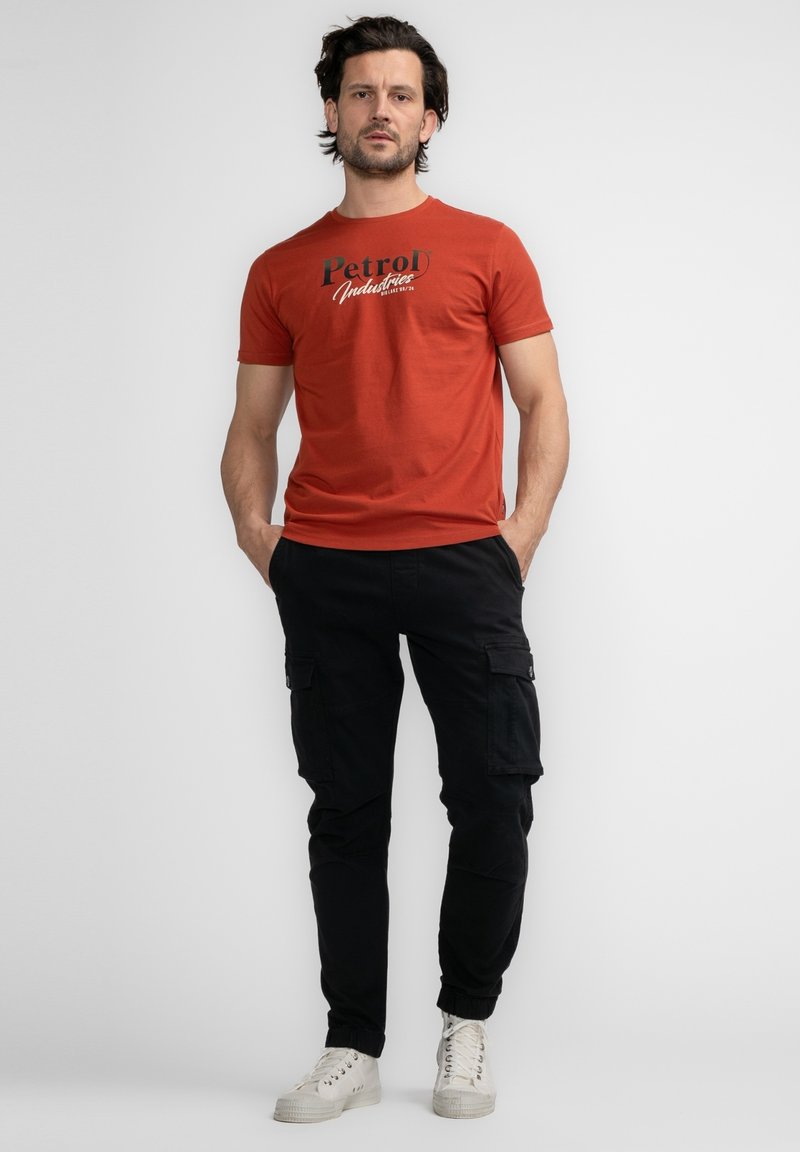 Oranje katoenen T-shirt met zwart bedrukte logo, gecombineerd met zwarte cargobroek met zakken, en witte veterschoenen.