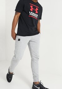 Camiseta negra con el logo rojo en negrita de "Under Armour", combinada con joggers gris claro que presentan una pequeña etiqueta de la marca, complementados con zapatillas deportivas negras.