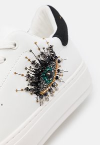 Vit sneaker med ett pärlbroderat öga i grönt, svart och guld, med intrikata dekorationer och en hälflik i svart mocka.