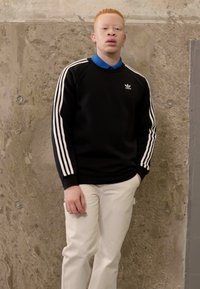 Sweat-shirt noir avec des rayures blanches sur les manches et un logo. Pantalons blancs. En arrière-plan, un mur gris texturé. Le modèle se tient les mains dans les poches.
