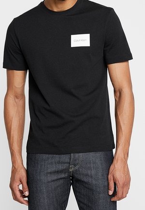 Černé bavlněné tričko s krátkým rukávem a bílou obdélníkovou nášivkou s nápisem „Calvin Klein“. Jednoduchý design s kulatým výstřihem.