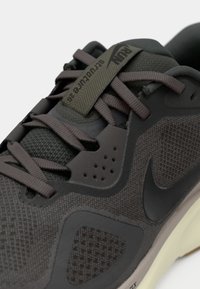 Scarpa da corsa Nike grigia scura con tomaia in mesh, lacci spessi, etichetta marchiata sulla linguetta e suola chiara vista da vicino.
